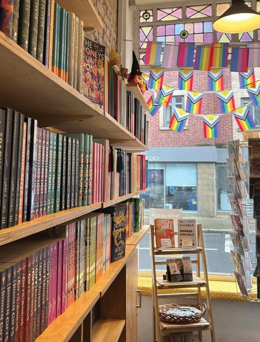 The Old Queeriosity Shop interior: colour-arranged bookshelves on the left, a wooden Queer+There zine A-frame in the foreground, Progress Pride bunting strung across the upper window, the Bretonside brickwork visible beyond.