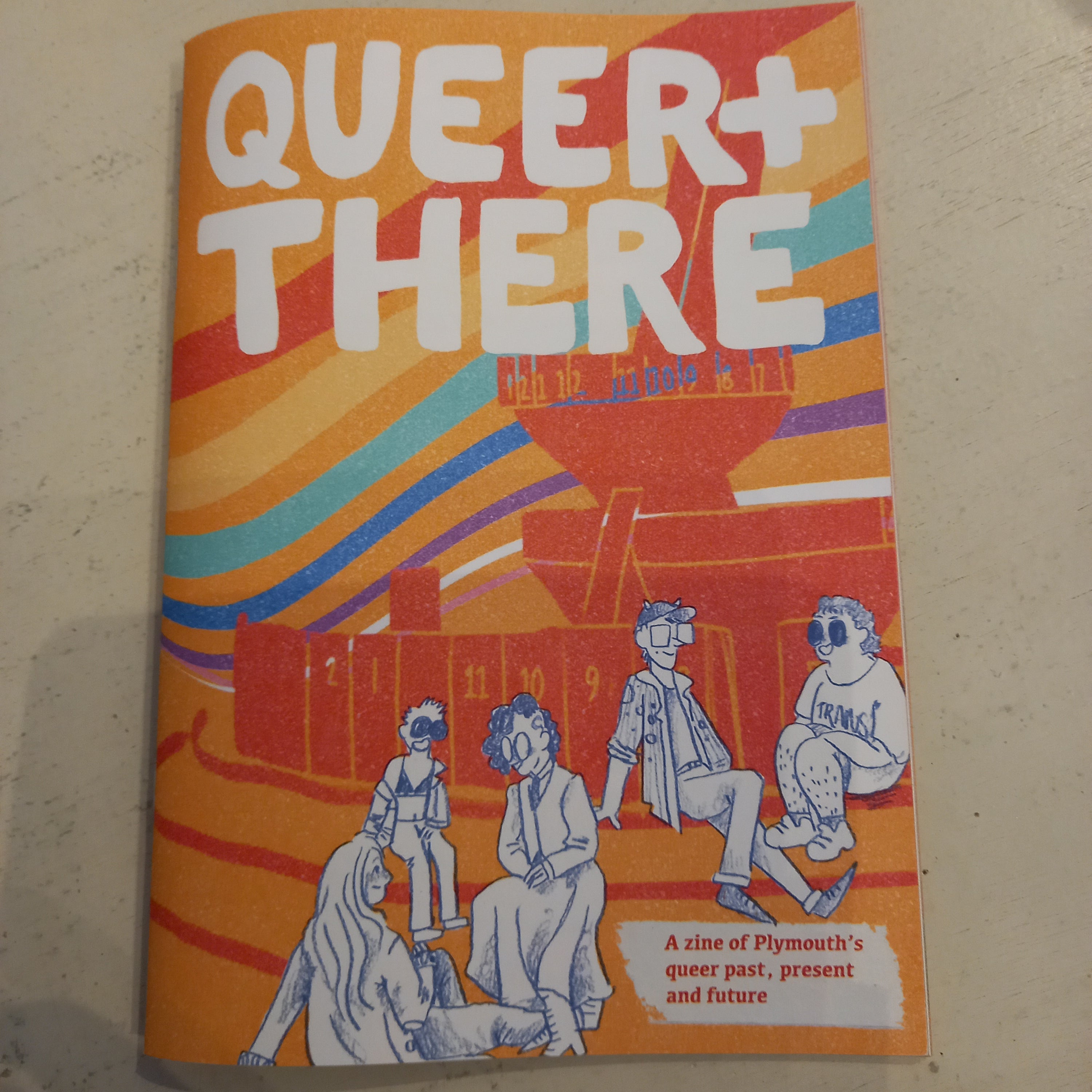 The Queer+There zine cover: an orange-and-rainbow striped background, five illustrated figures on a red harbour-defence boat, with one figure wearing a TRANS t-shirt.
